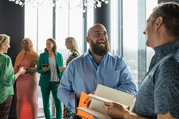 Two white men both wearing blue shirts chat standing in an office setting with five women and a man chatting in the background